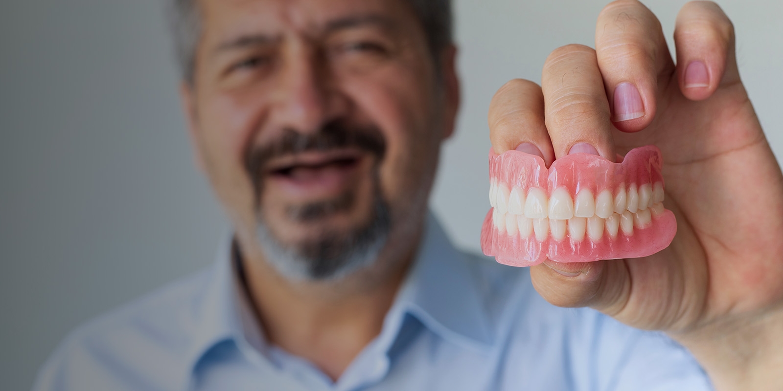 An older man holding dentures toward the camera.